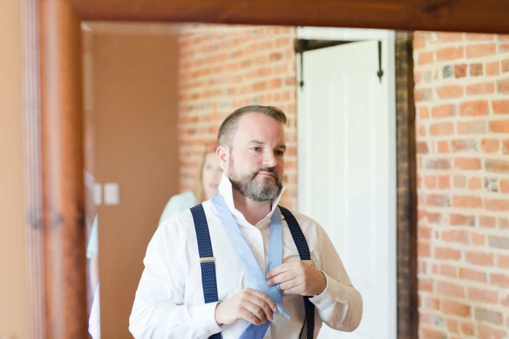 Person adjusting suspenders while getting dressed in room with exposed brick wall.