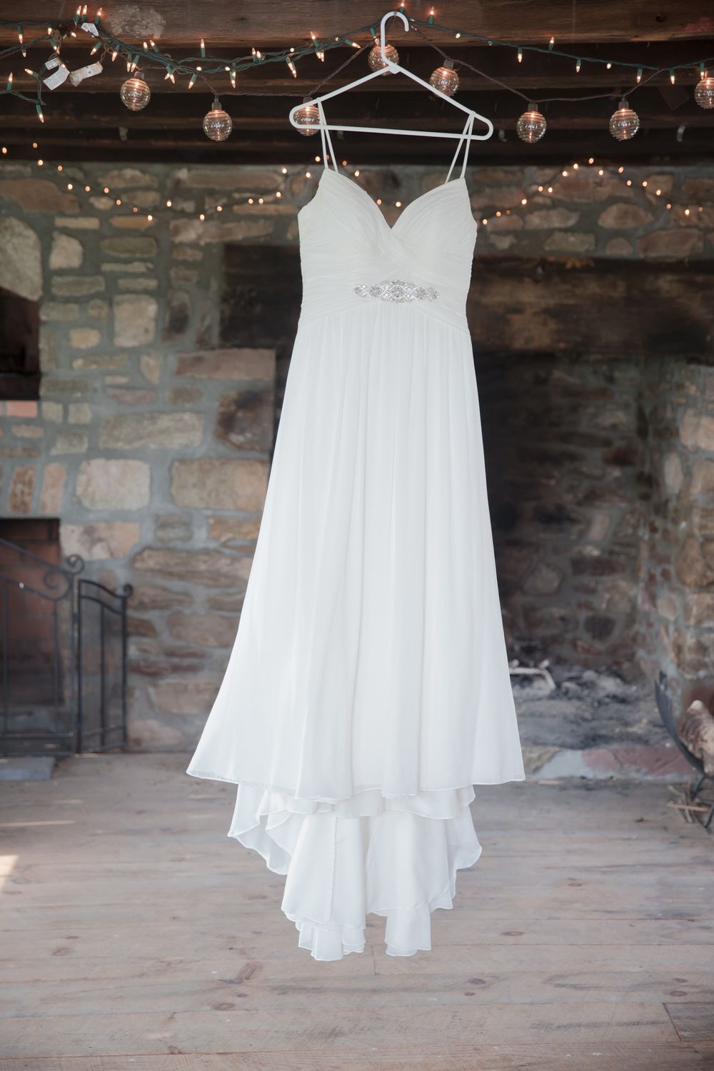 White wedding dress hanging on display with string lights in rustic stone room setting.