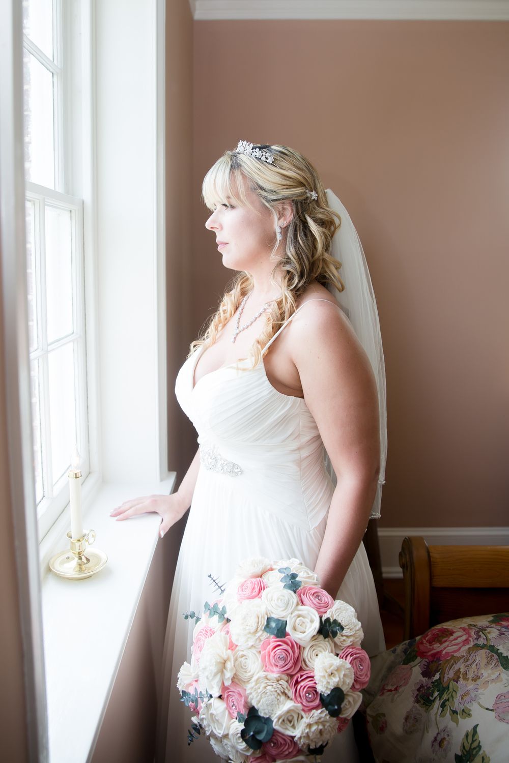 Bride in white dress holding pink and white rose bouquet while looking out window.