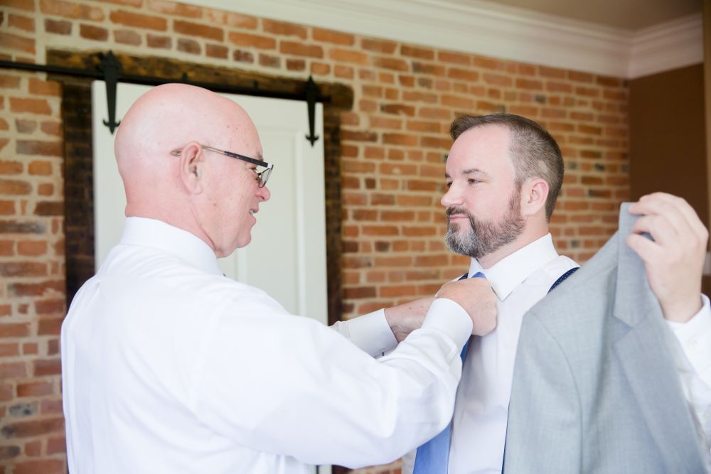 Groomsmen helping each other prepare with formal attire adjustments in brick room.