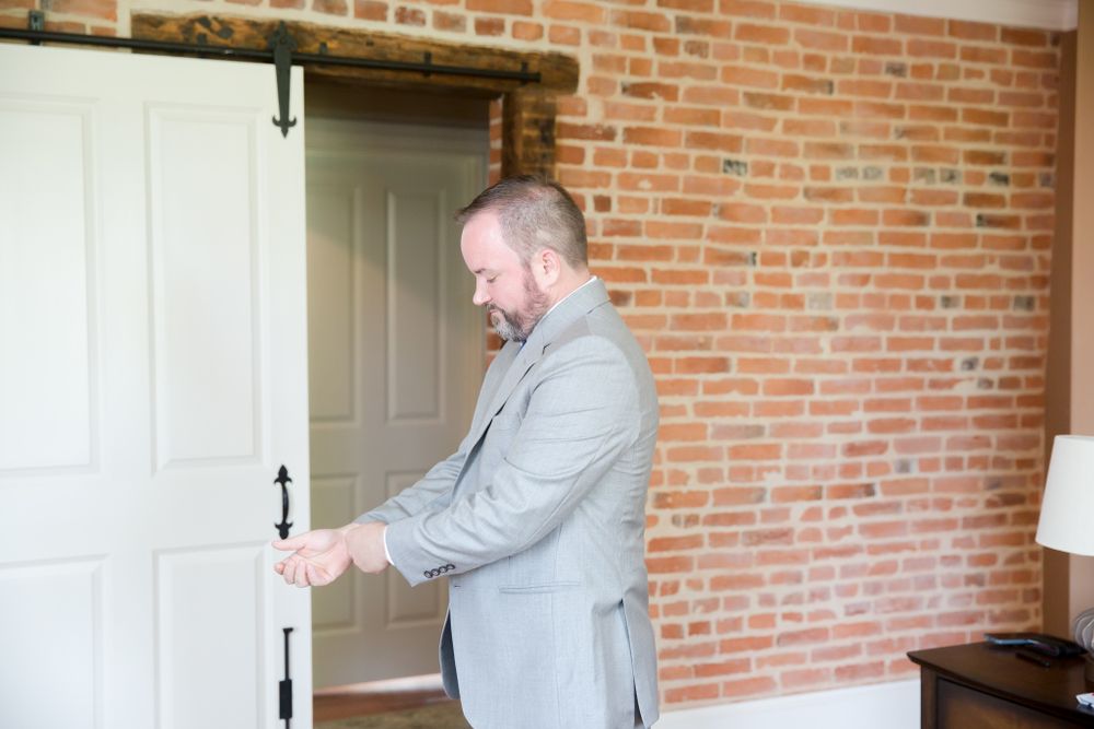 Groom adjusting sleeve in grey suit against exposed brick wall.