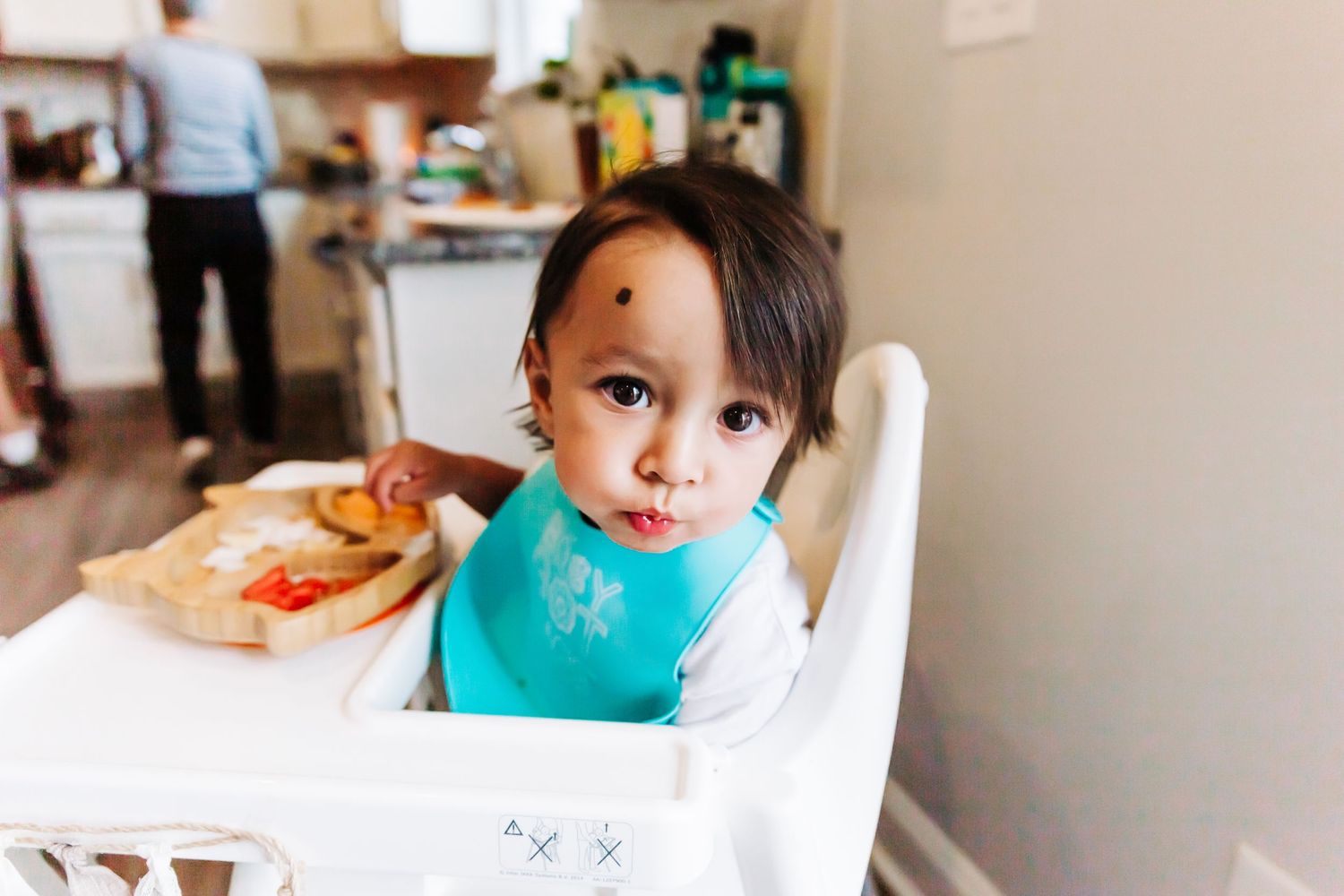 A one year old boy looking into the camera while he eats
