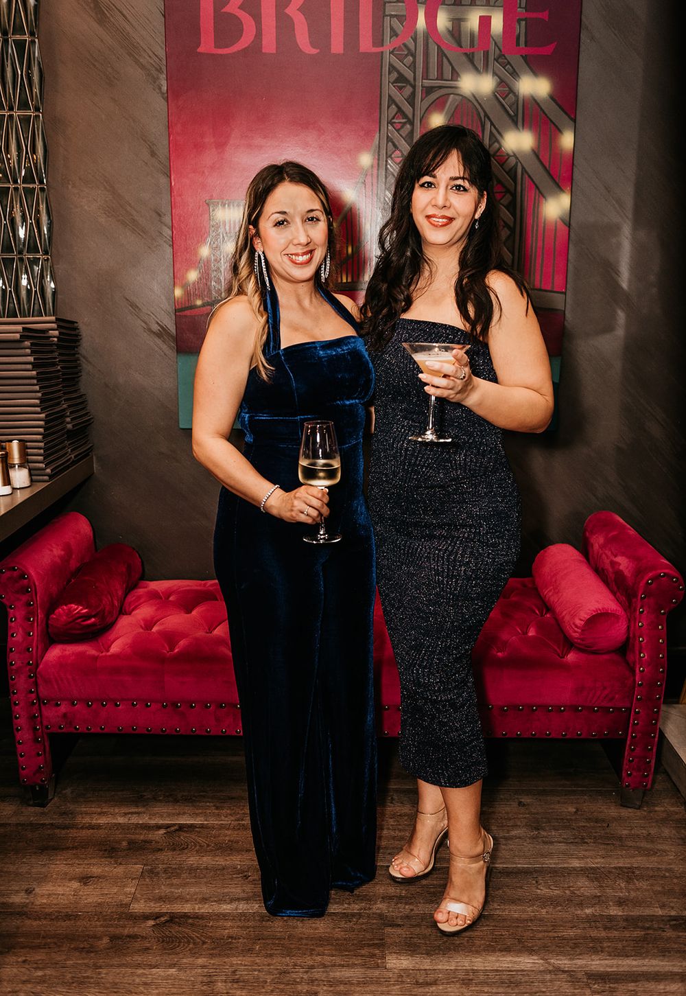 Two women in elegant evening wear pose with cocktails against a red backdrop with plush velvet couches.