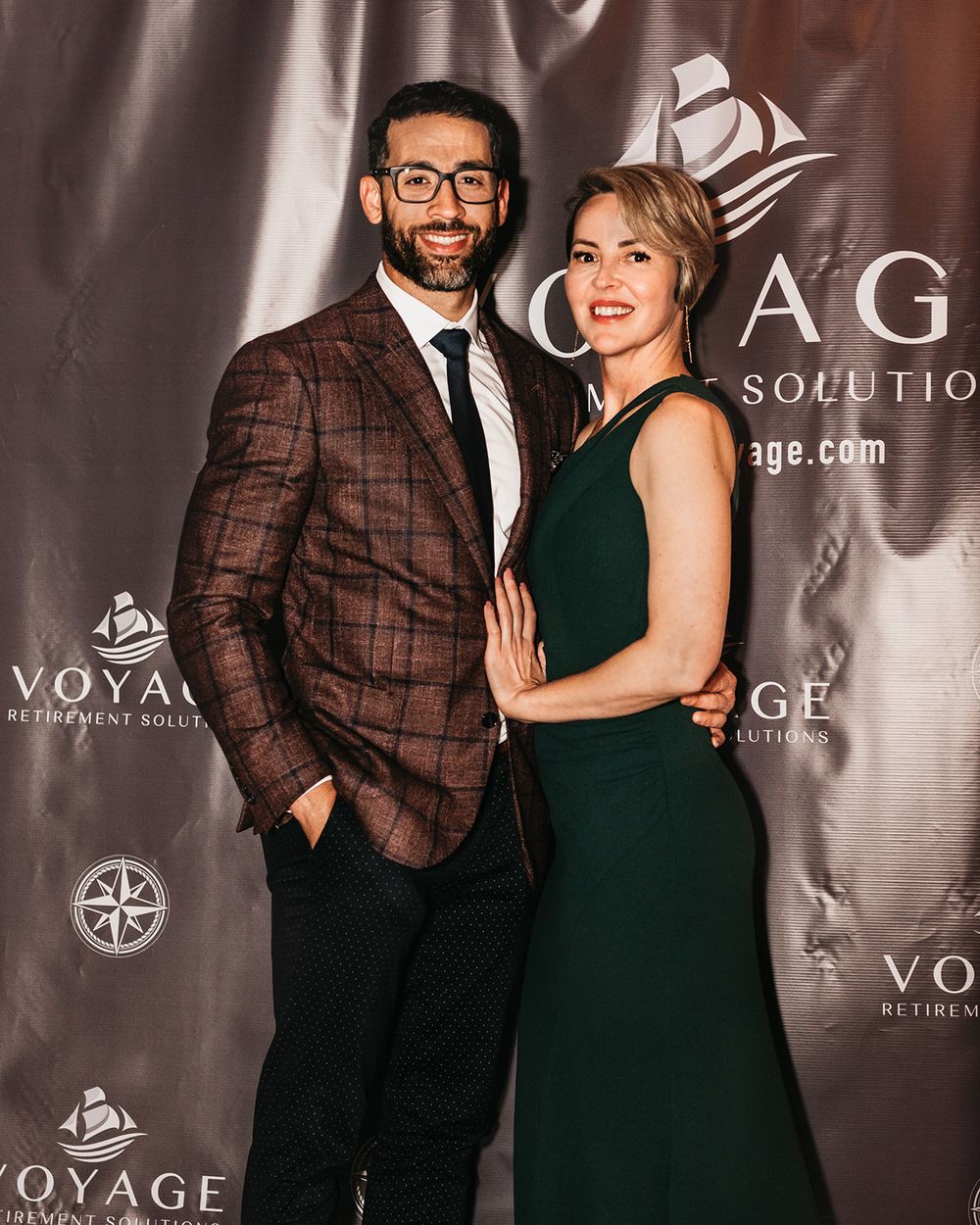 A couple poses against a branded backdrop wearing formal evening attire.