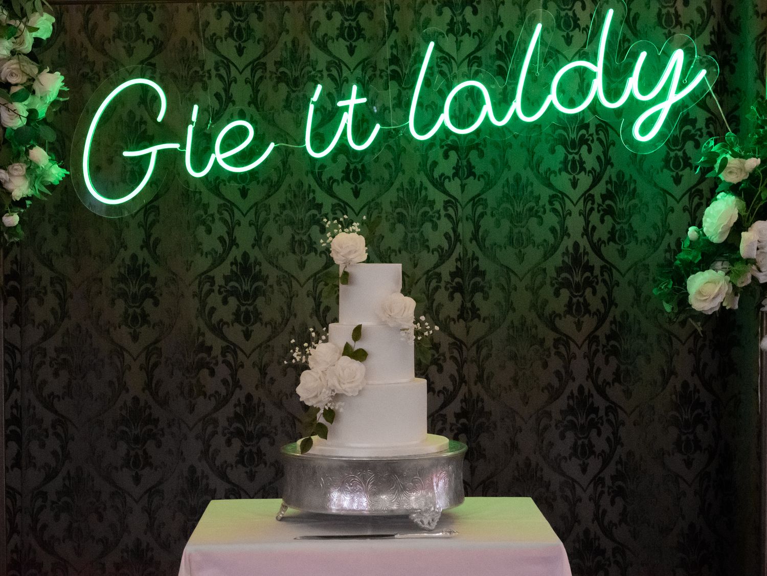 Green neon sign reading 'Gie it laldy' above a white wedding cake.