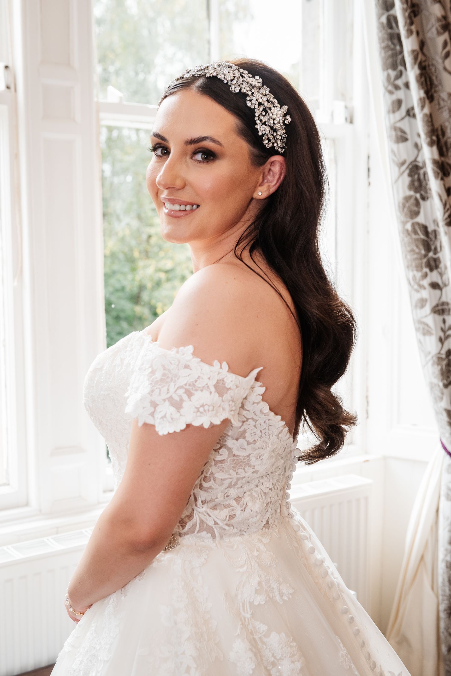 Elegant bridal portrait showing lace wedding dress and crystal headpiece near window.