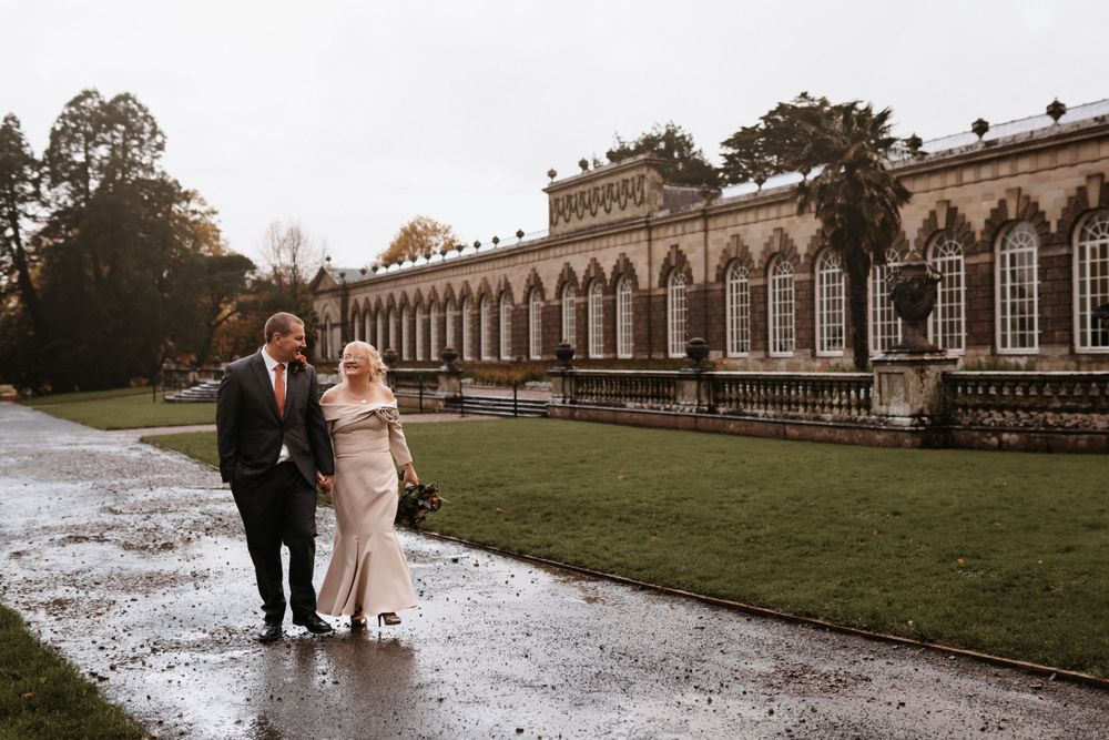 Newlyweds walk along historic building exterior on rainy day at The Orangery Margam Park