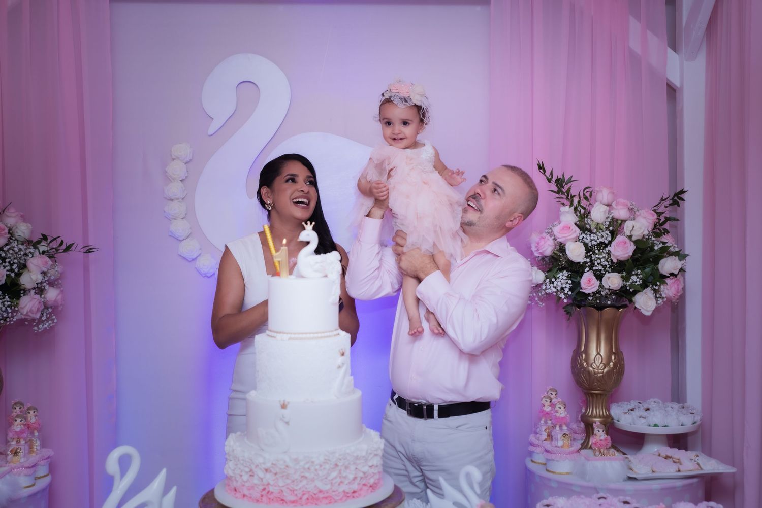 A couple celebrates at a beautifully decorated pink and white themed party with a swan backdrop and floral arrangements.