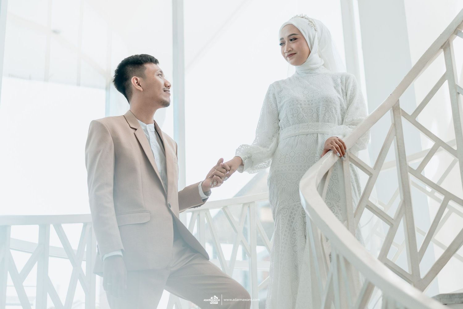 Elegant Muslim couple in white and beige formal attire pose on modern white staircase.