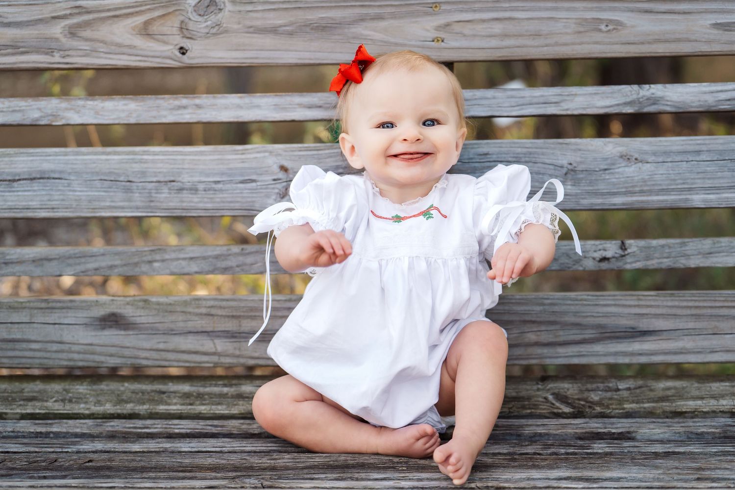 infant girl wearing a white bubble and a red bow in her hair smiling sitting on a bench at Phil Hardberger Park