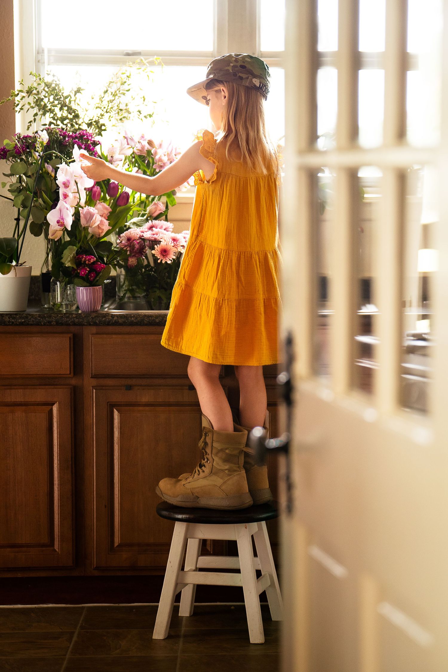 through a door a little girl in  yellow sundress, her dad's boots & military cap stands on a step stool to reach flowers