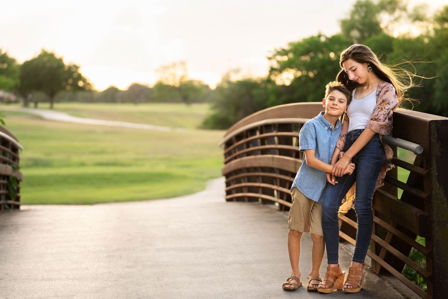 a teen girl leans against the railing of a bridge and looks down at her younger brother whose hand she is holding