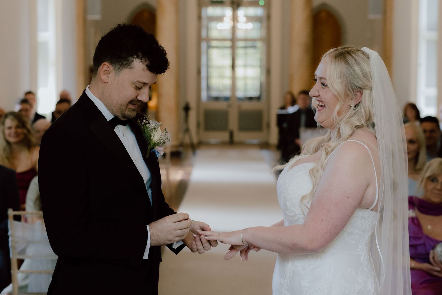 An indoor wedding ceremony showing a ring exchange with guests in the background.