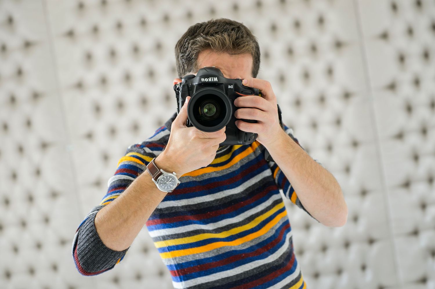 Someone wearing a striped shirt takes a photo with a professional DSLR camera against a patterned background.