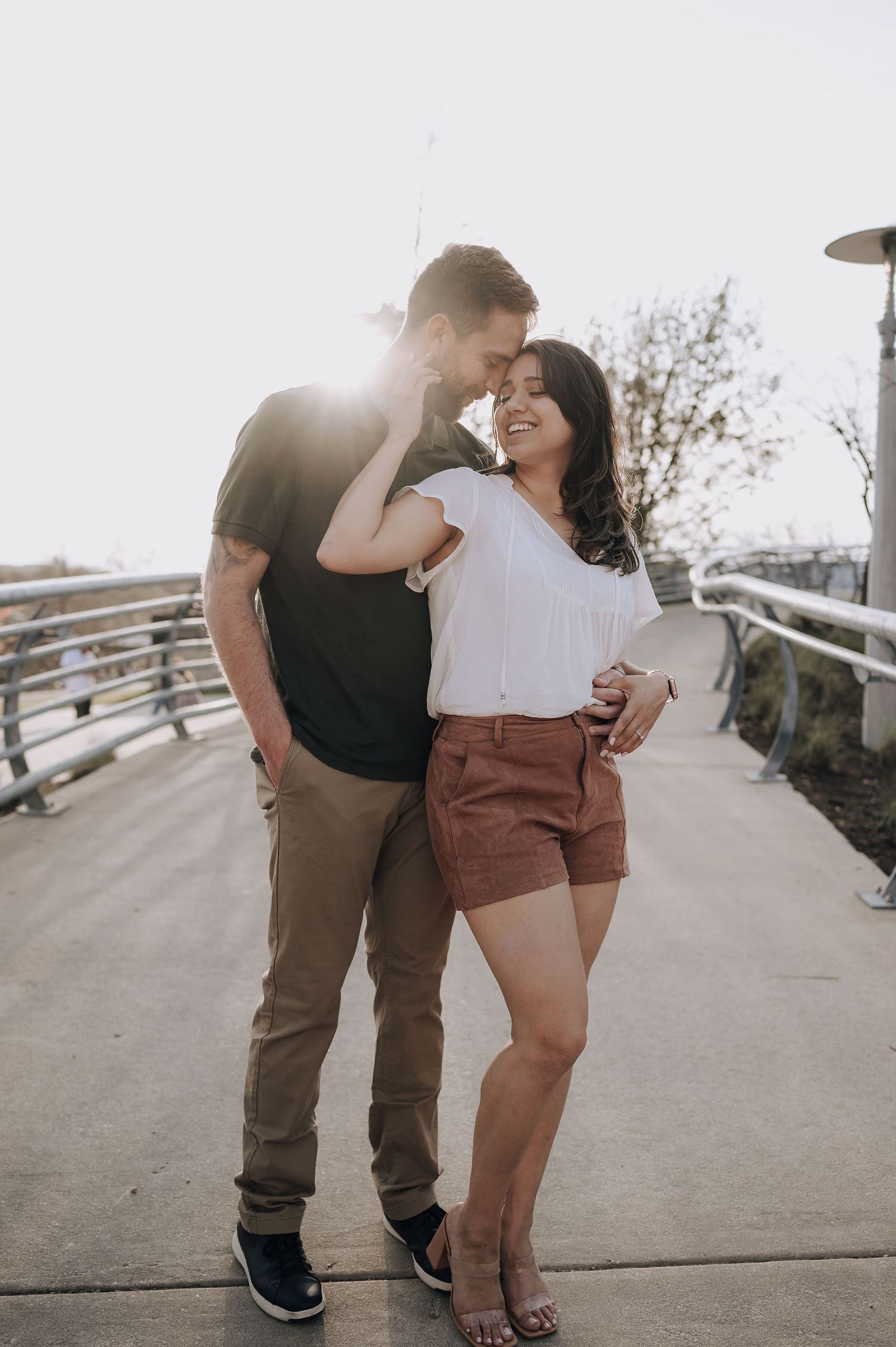 A sunlit couple shares a romantic moment on an elevated walkway with metal railings.