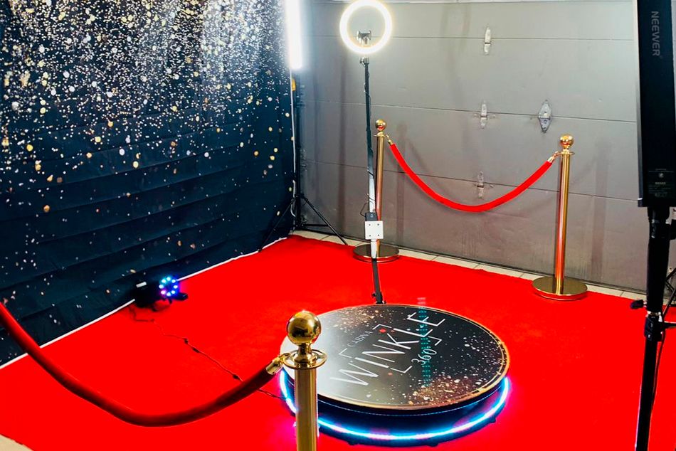 360 photo booth setup with ring light, red carpet, and gold stanchions against a dark backdrop.