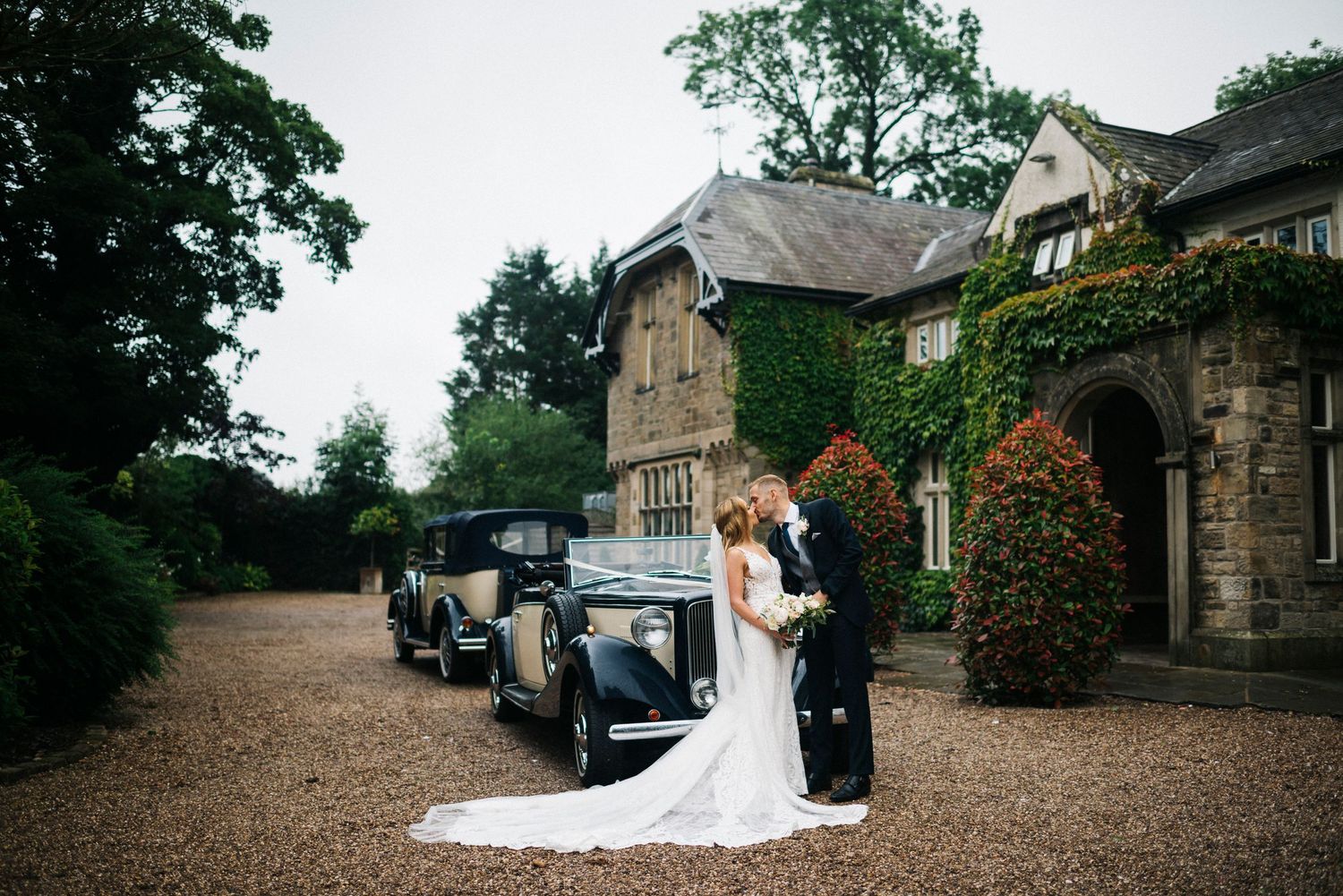 Vintage wedding scene with classic car parked outside stone cottage on gravel driveway.