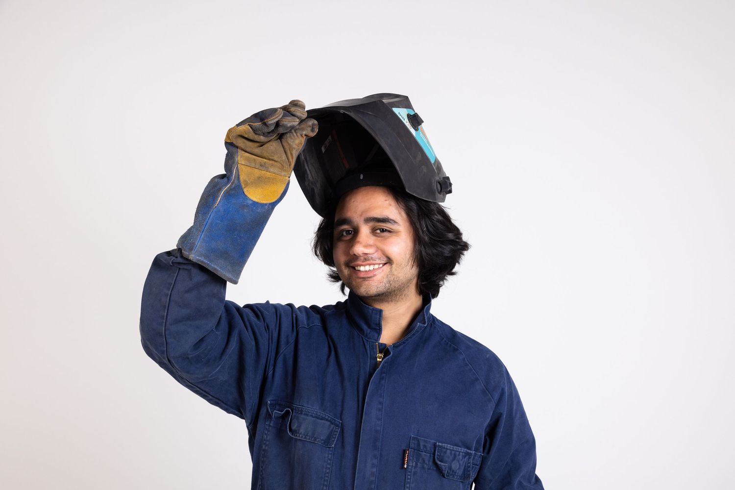 Trade worker in blue coveralls lifting welding mask against white background.