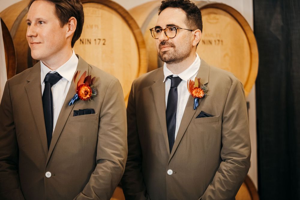 Groomsmen in matching brown suits with orange boutonnieres stand before wine barrels.