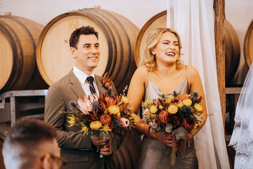 Wedding party members holding autumn-colored bouquets pose in front of wine barrels.