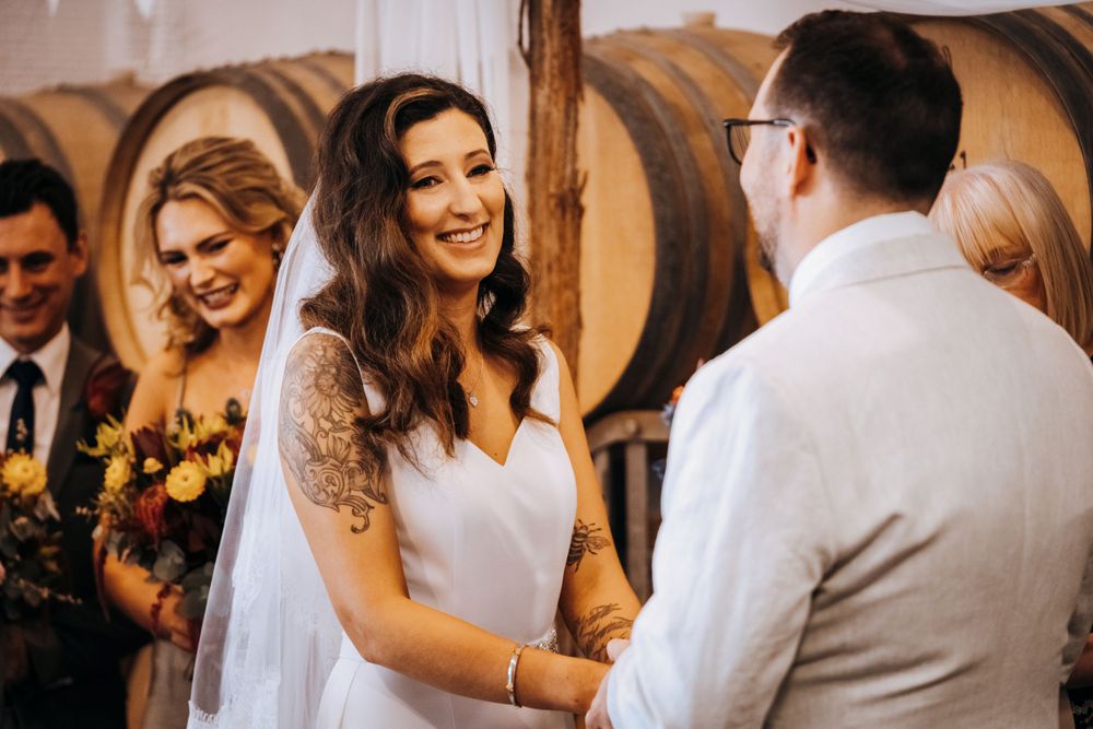 Bride and groom exchange vows during ceremony in wine cellar with wedding party looking on.