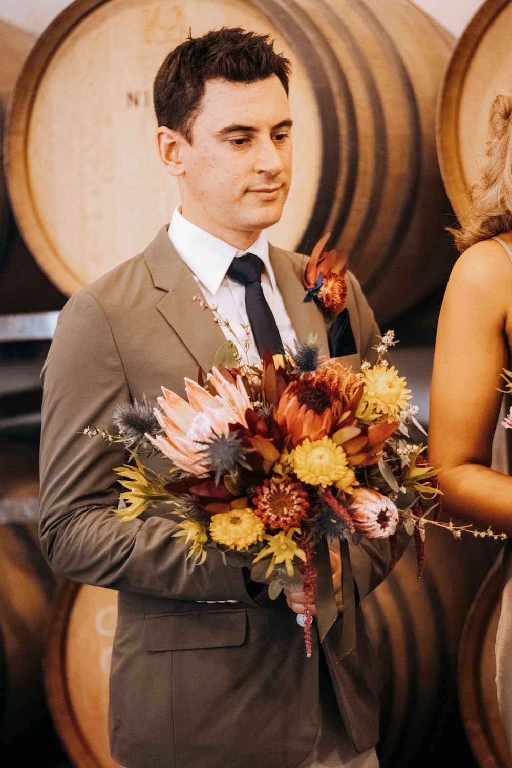 Groomsman in gray suit holding vibrant fall-colored wedding bouquet against wine barrel backdrop.