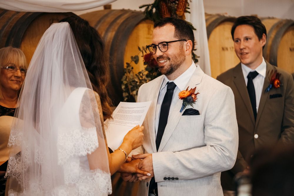 Bride and groom hold hands during wedding ceremony in rustic wine barrel room.