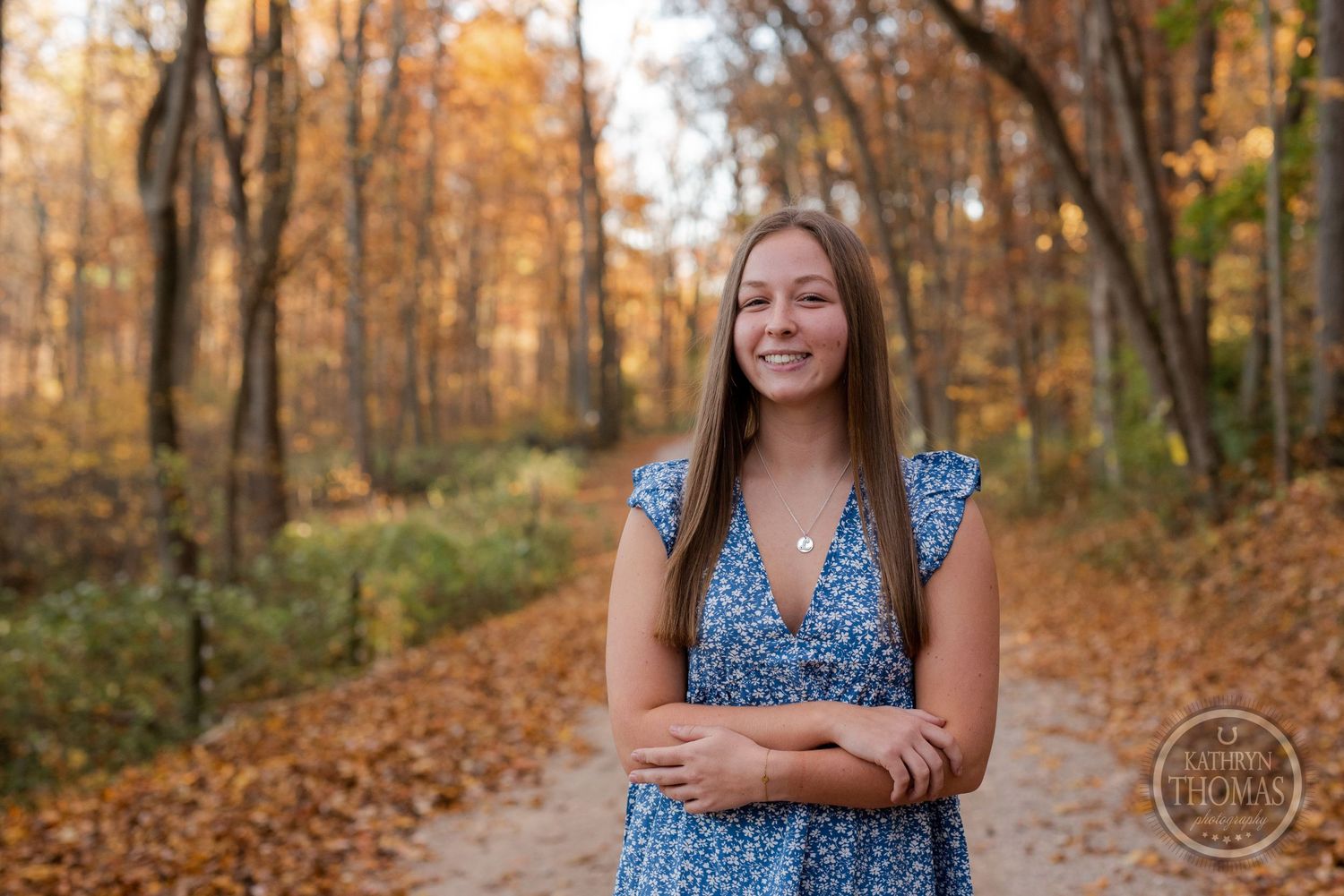 Someone wearing a blue floral dress smiles while standing on a forest path in autumn.