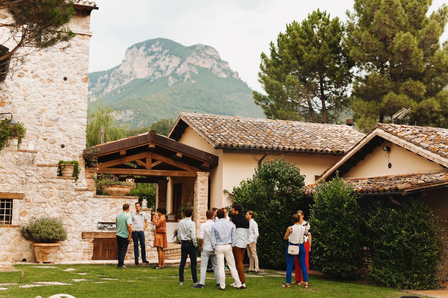 Small gathering of guests outside a rustic stone building with mountains in the background.