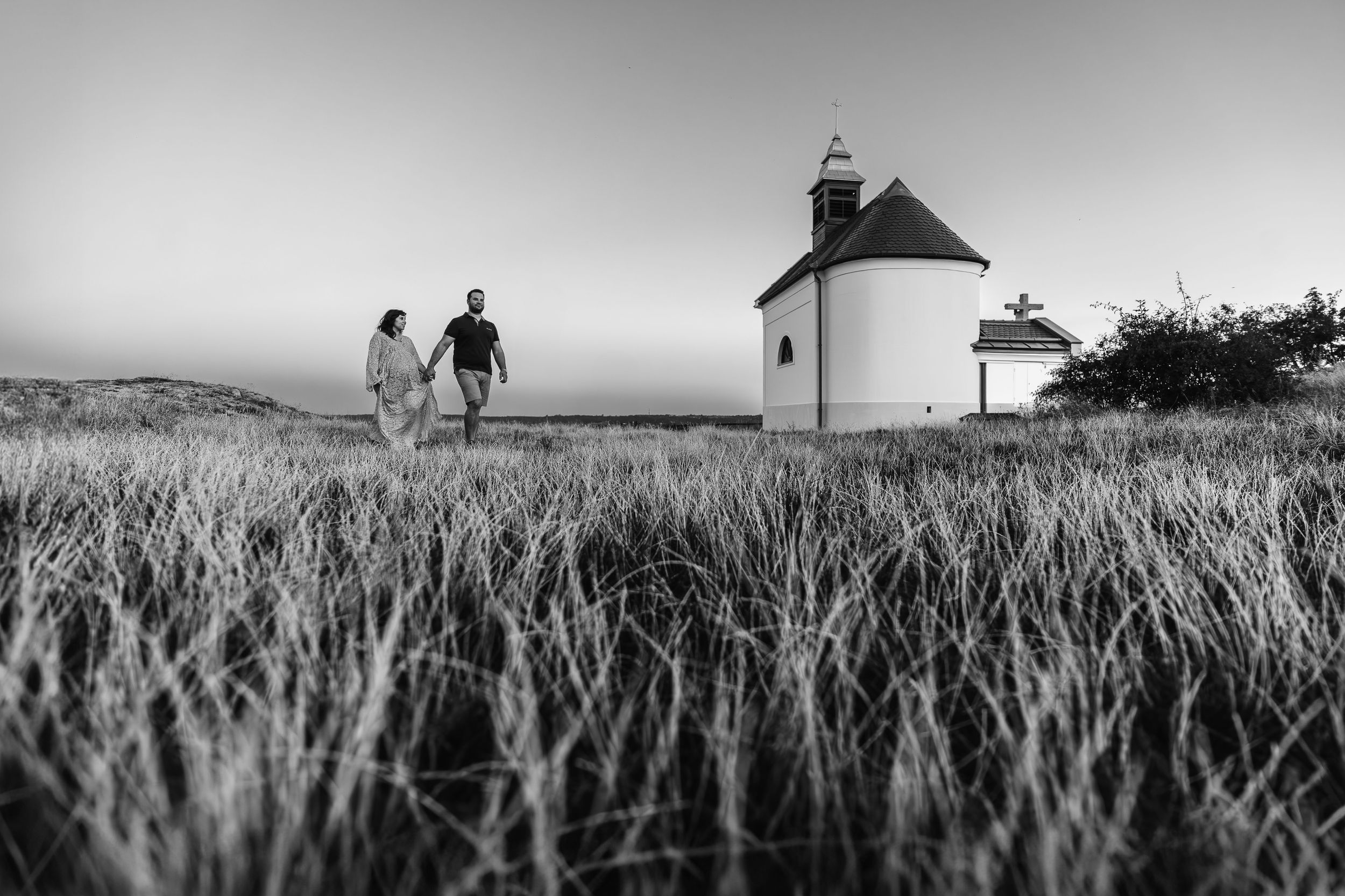 Two people walk towards a white lighthouse through tall grass in black and white landscape photography.