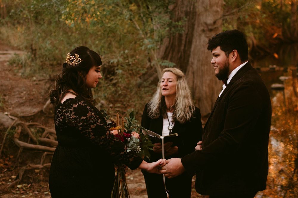 An intimate outdoor wedding ceremony takes place among tall trees in an autumn forest setting.
