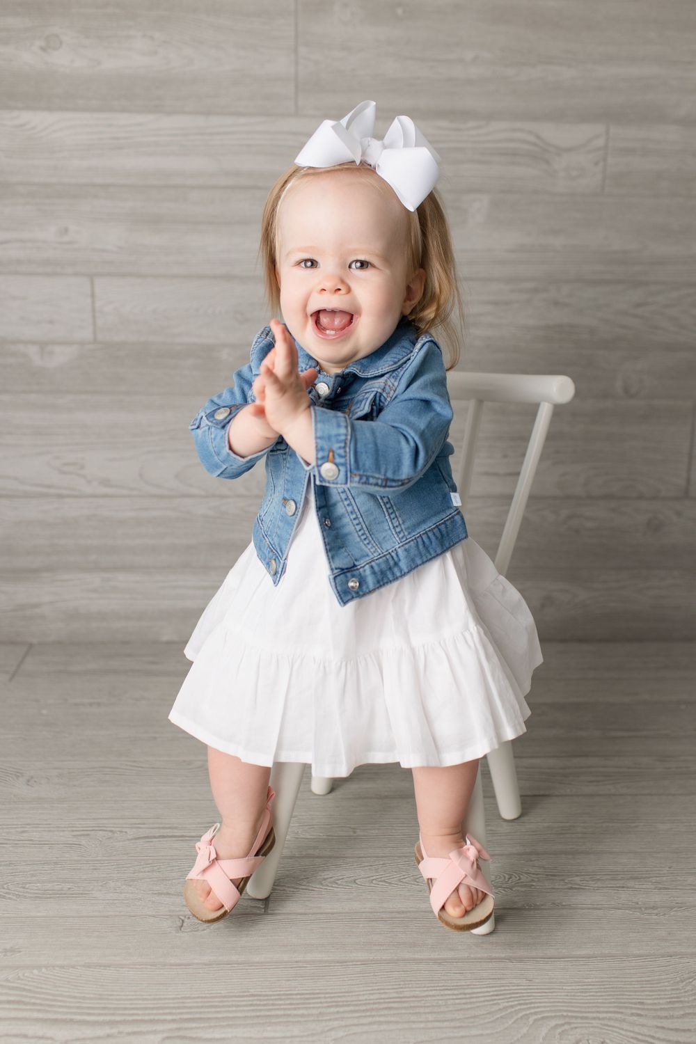 Happy toddler in white dress and denim jacket claps while standing by white chair.