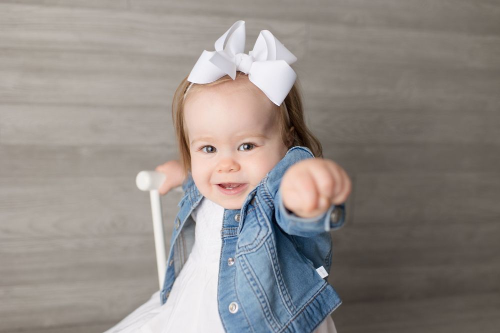 Toddler in white dress and denim jacket points forward while posing with white chair.