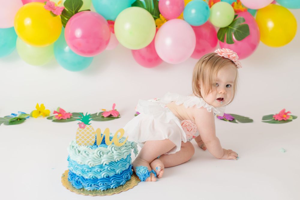 A festive cake smash setup with colorful balloons and a blue ombre birthday cake on white backdrop.