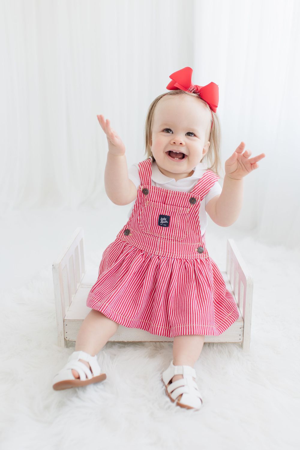 Happy toddler in red striped dress raises arms while sitting in white chair.