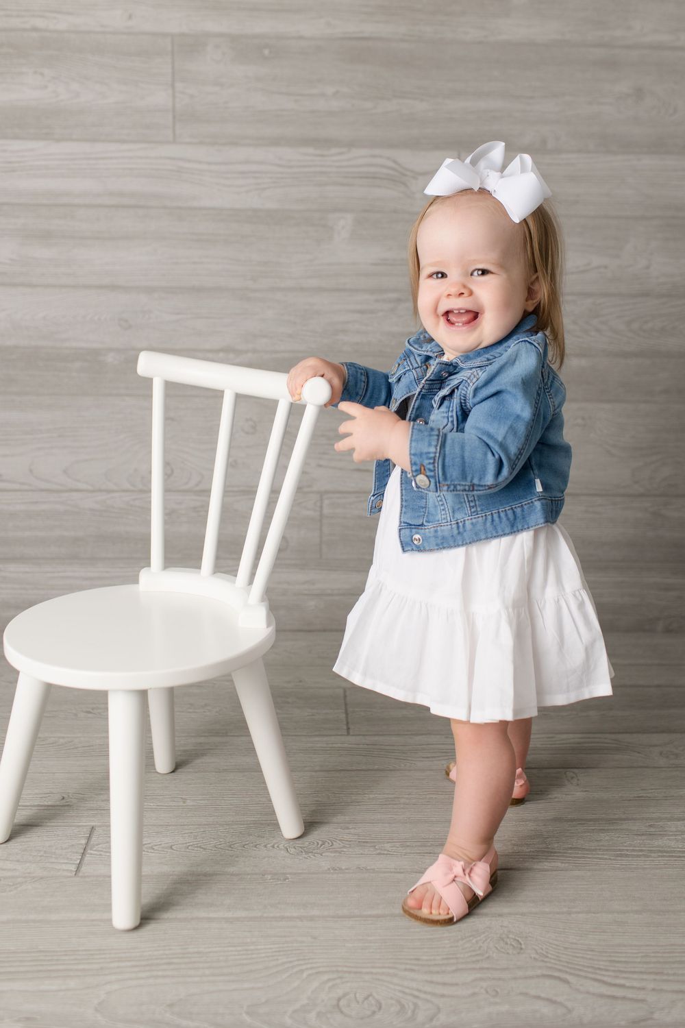 Toddler in white dress and denim jacket stands holding white chair with bow in hair.