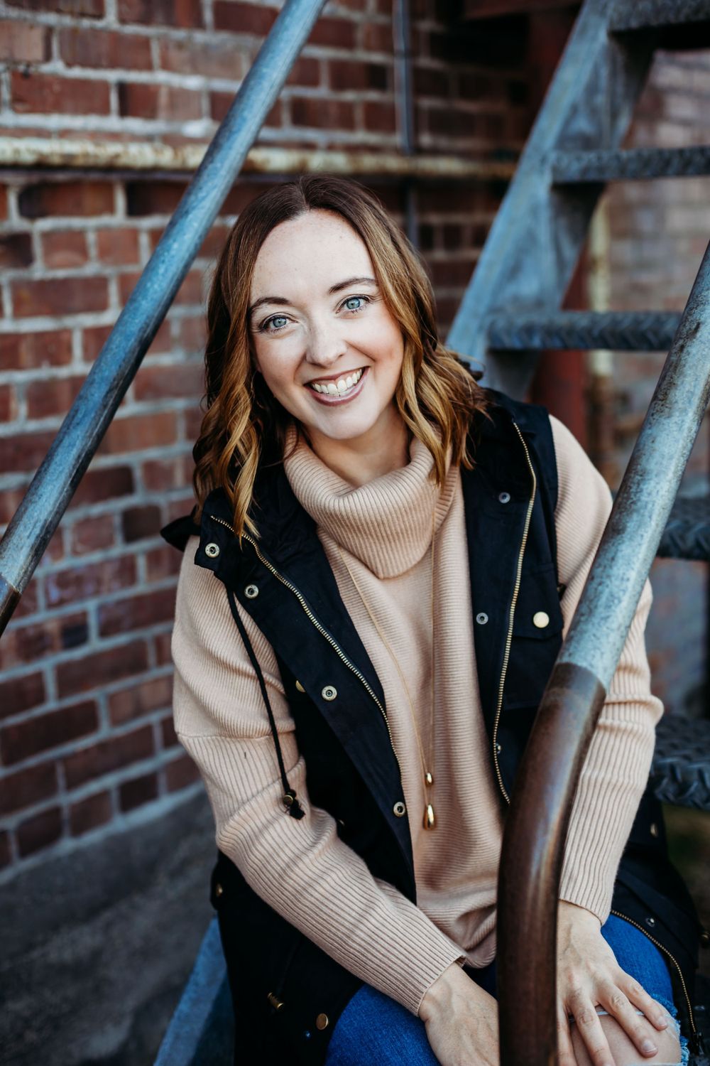 Casual portrait against brick wall wearing beige sweater and black vest.