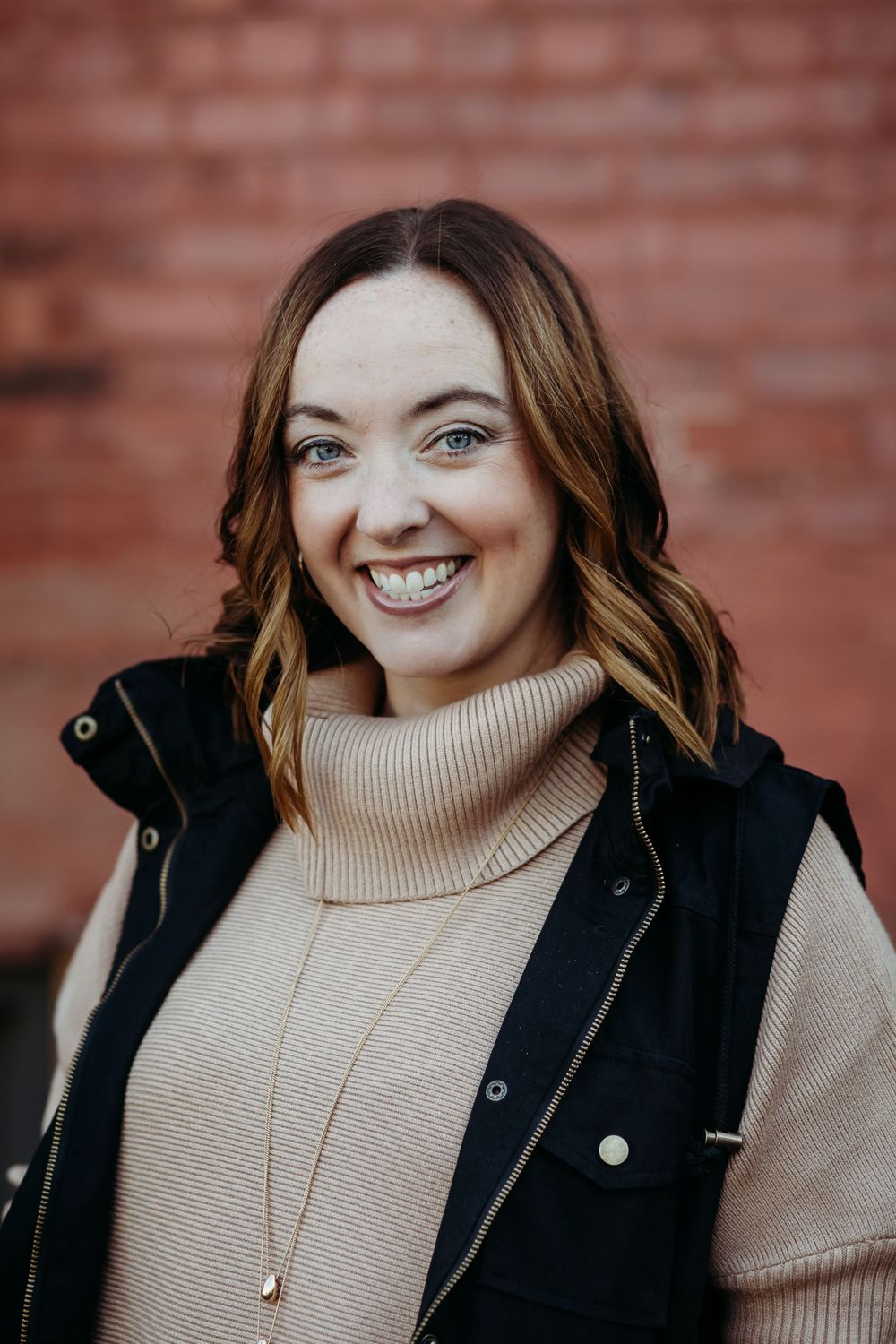 Portrait of someone in beige turtleneck and black vest smiling against red brick background.