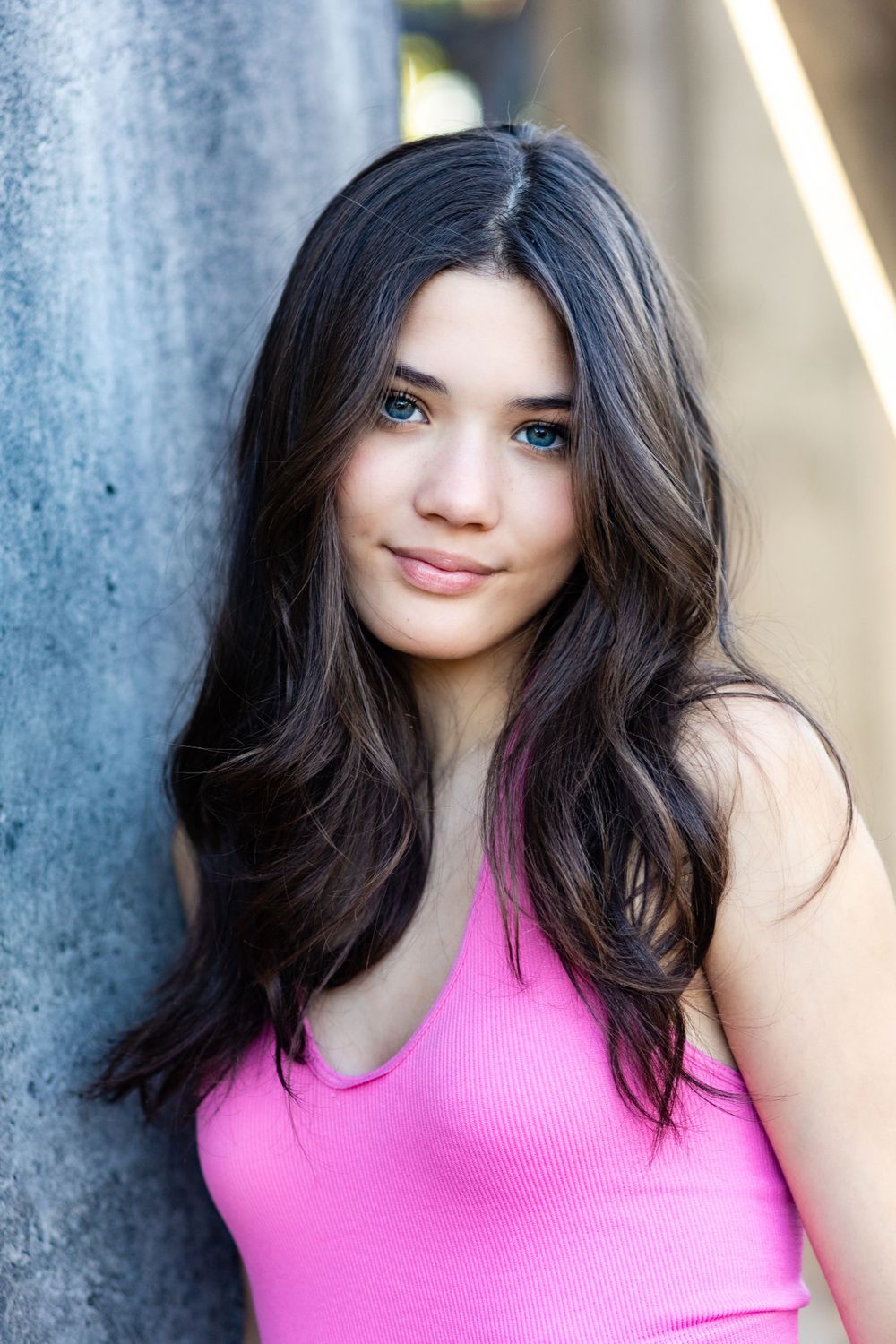 Portrait of someone in bright pink tank top leaning against pillar with long dark hair.