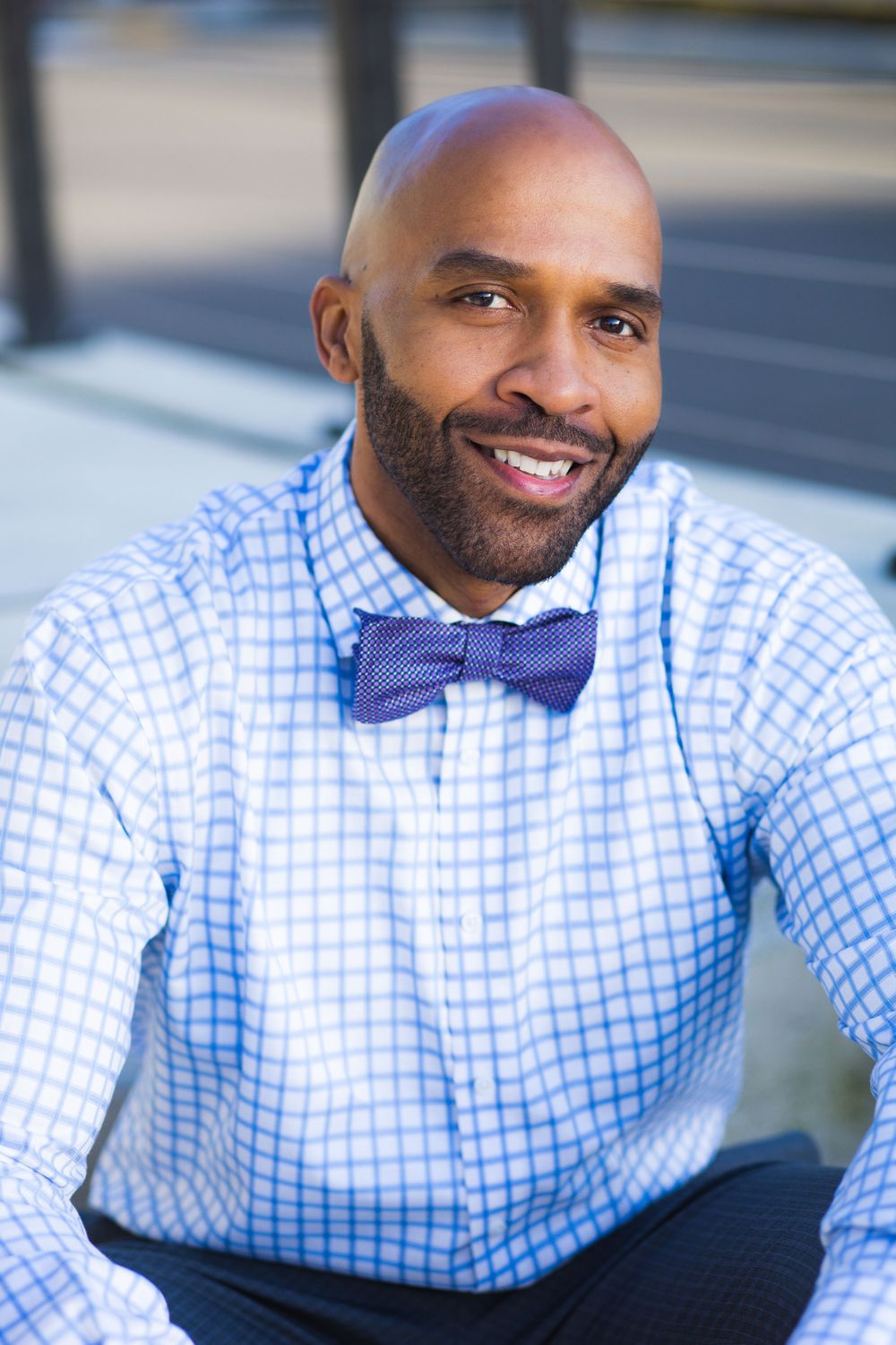Professional portrait of a person in a light blue checkered dress shirt and purple bow tie smiling warmly.