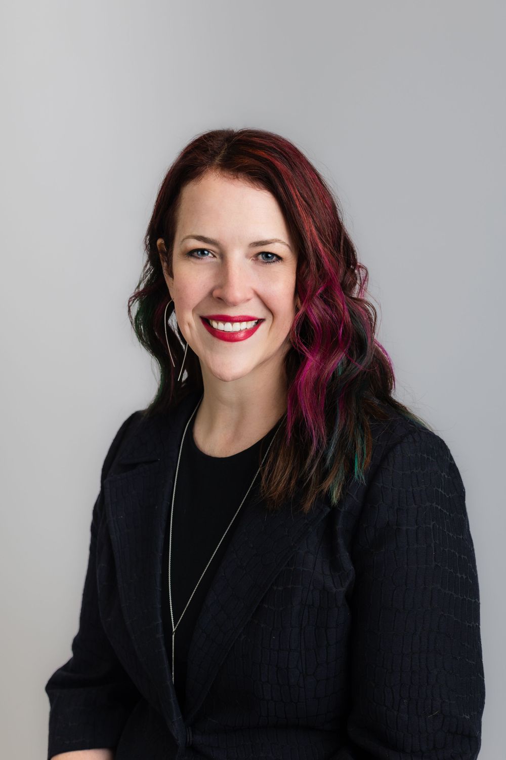Professional headshot of woman with vibrant pink-streaked hair wearing black blazer against grey background.