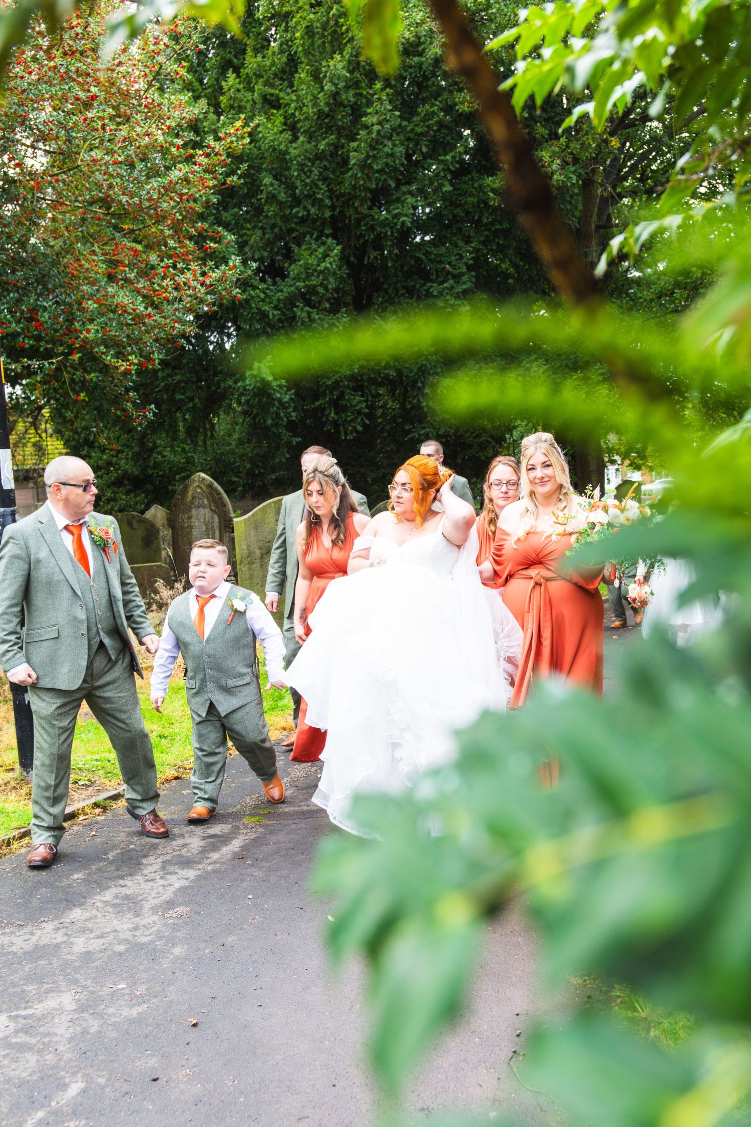 A wedding party walks down a tree-lined path with bridesmaids in coral dresses.