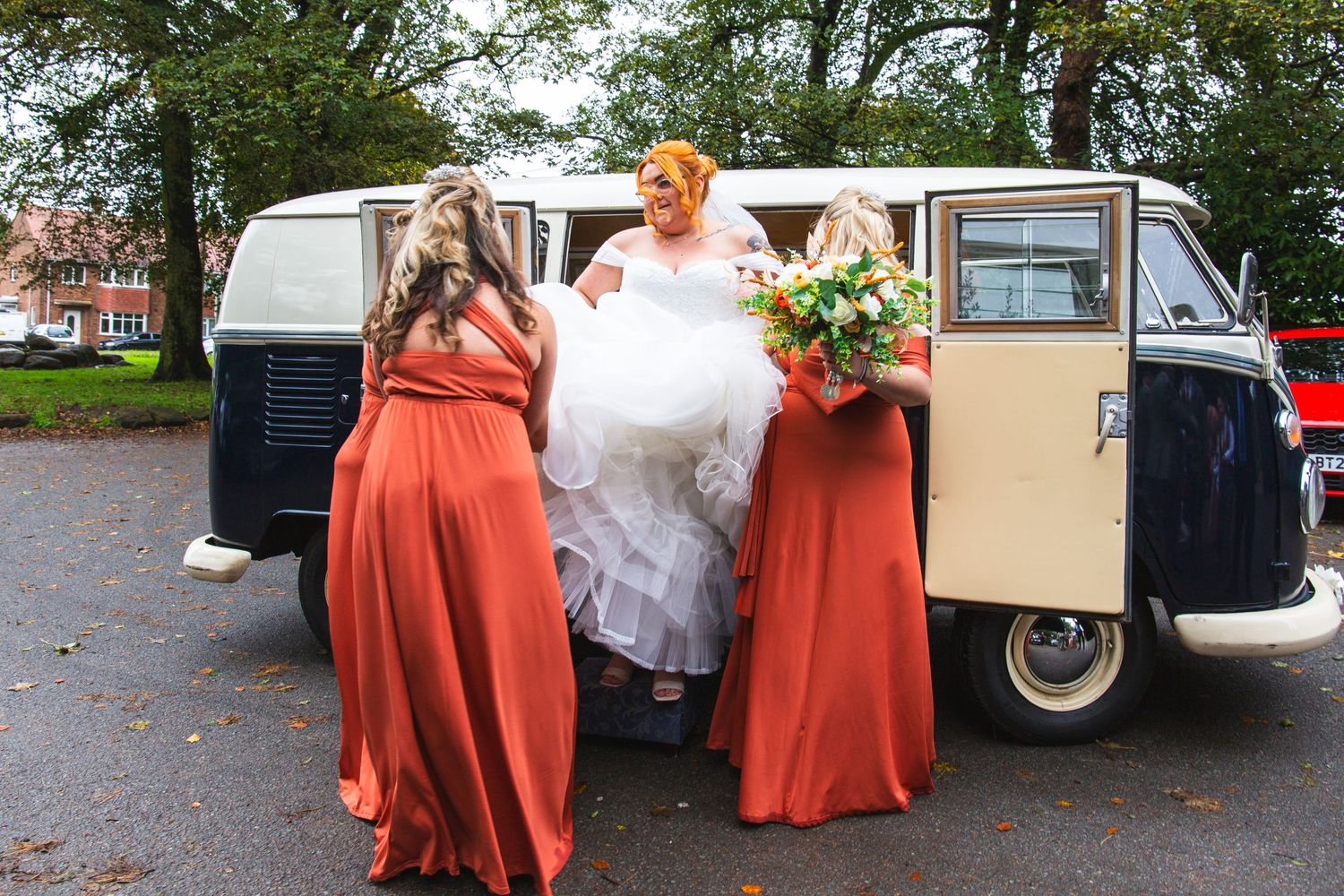 Wedding party helping with dress near a vintage VW van in coral-colored dresses.