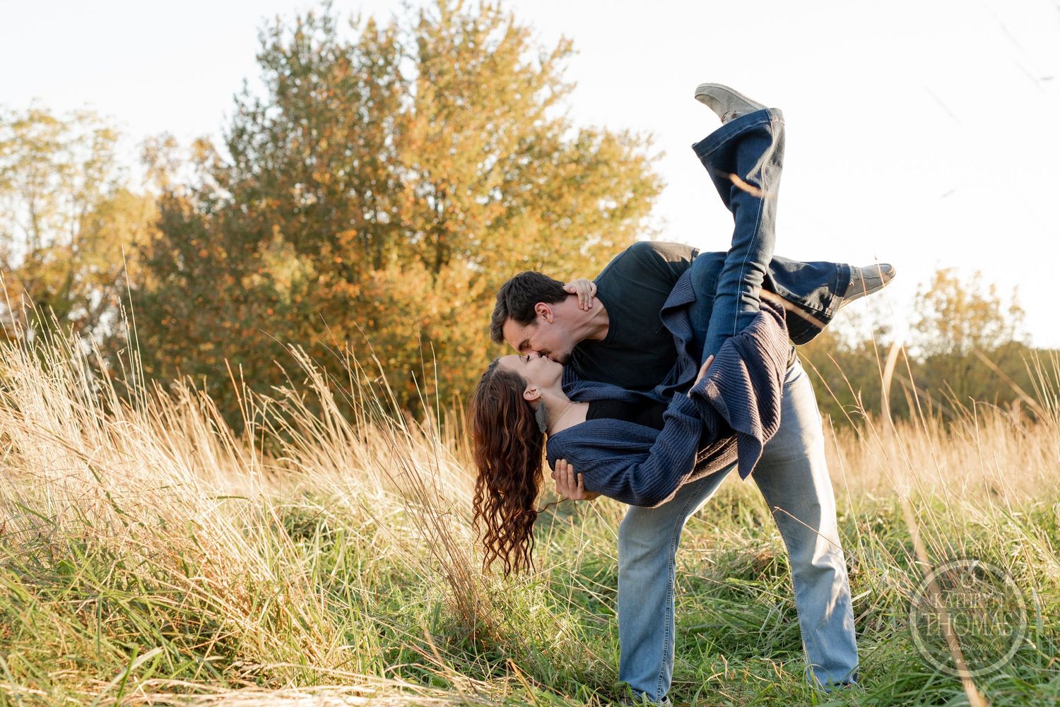 A playful dip pose in a grassy field during golden hour with autumn trees in the background.