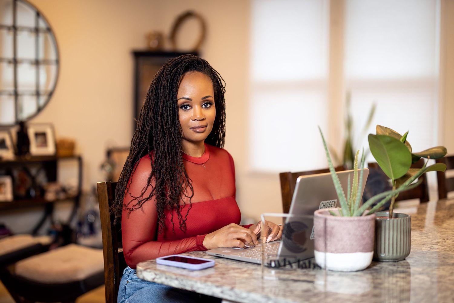 A person in a red sweater works on a laptop at a desk decorated with plants and home decor.