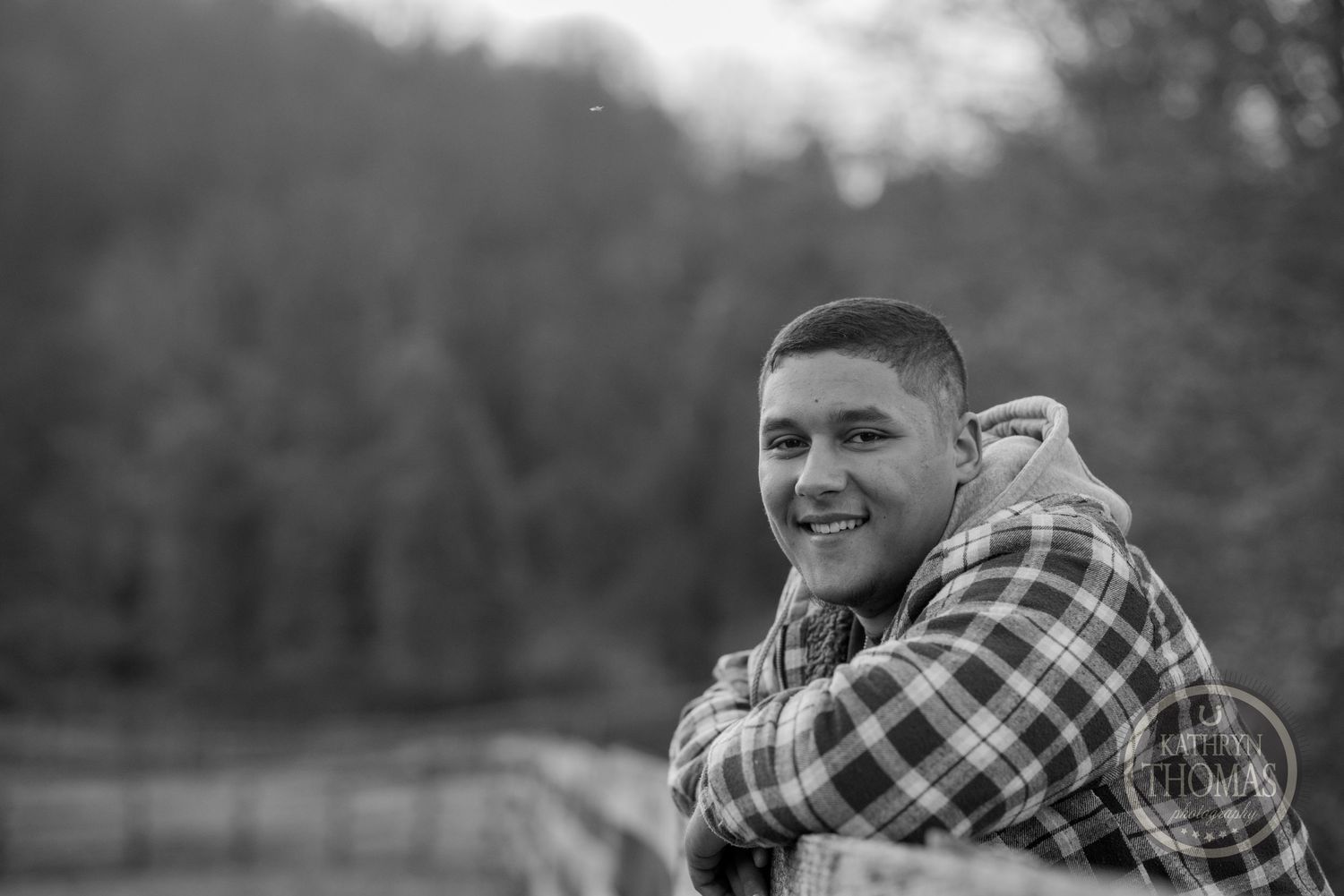 Black and white outdoor portrait of someone in a plaid shirt smiling against a blurred natural background.