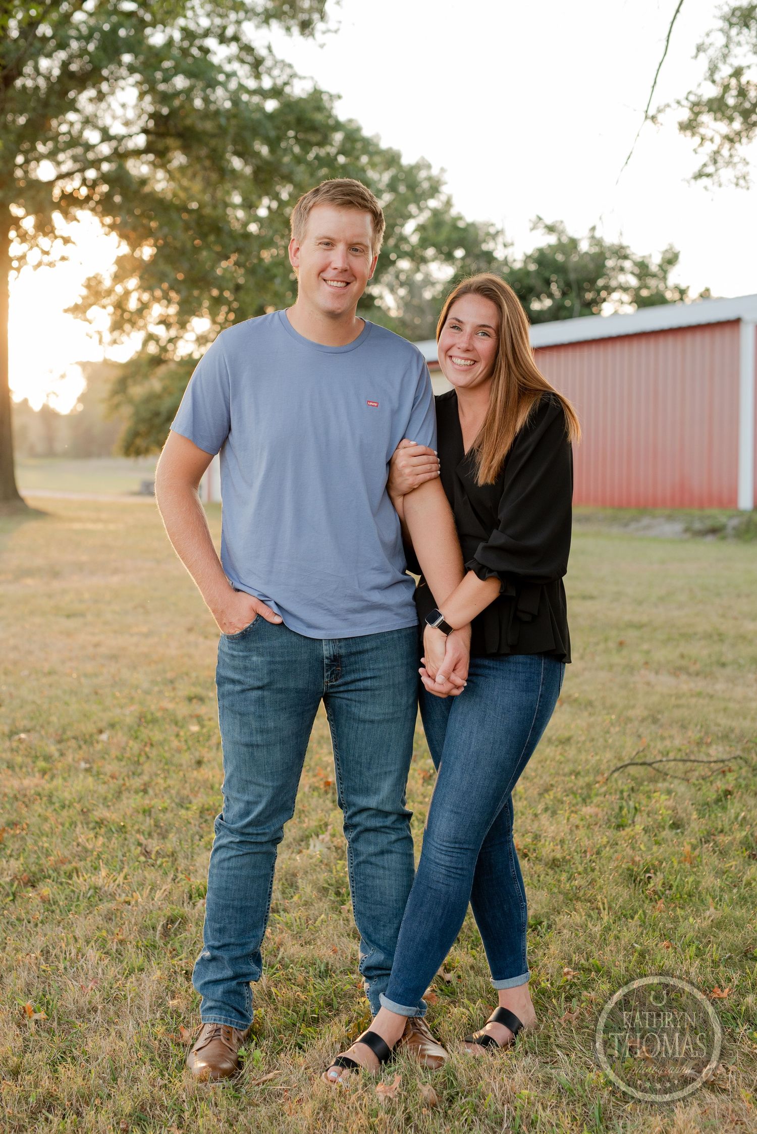 Couple posing together in casual wear with red barn and sunset in background.