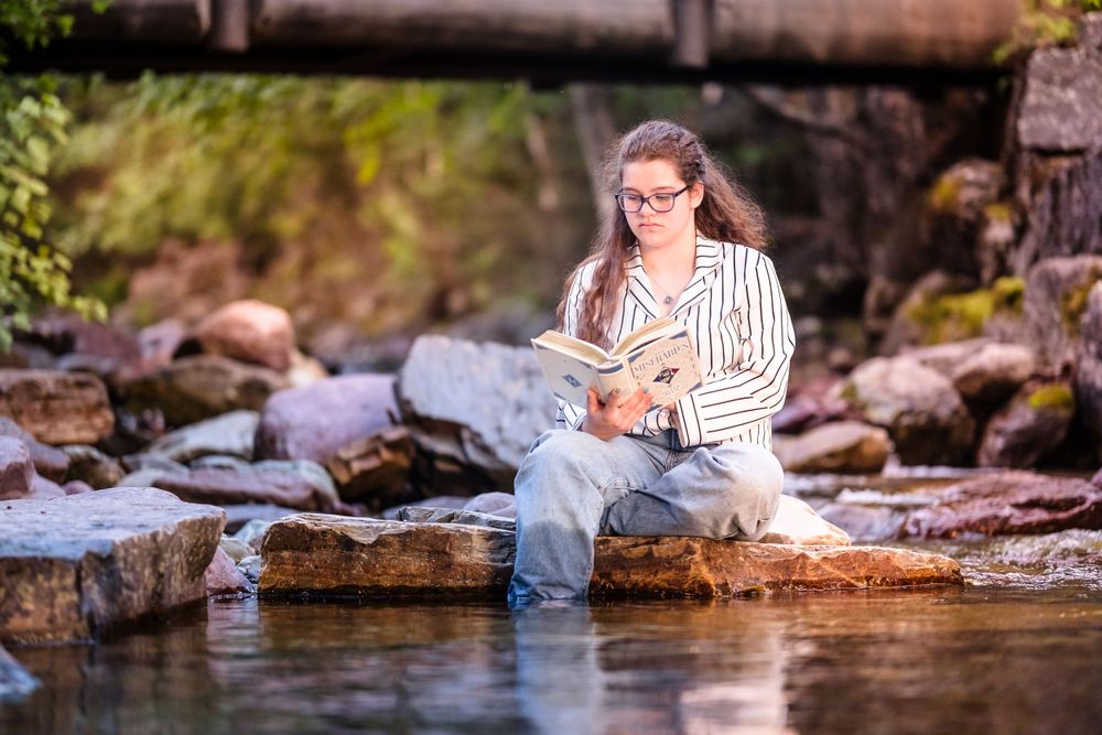 Person in striped shirt relaxing by a stream while reading on rocky shore.