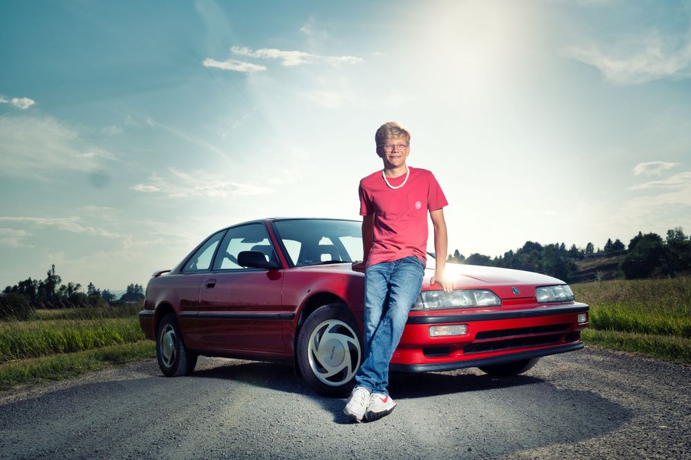Person in red shirt poses leaning against maroon sedan on rural road at sunset.