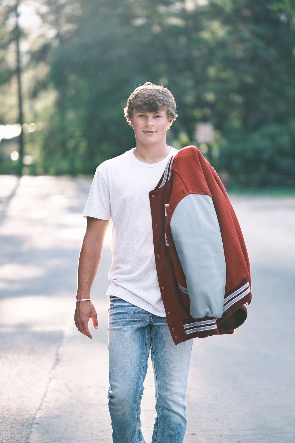 Casual portrait of student carrying varsity jacket on tree-lined path.